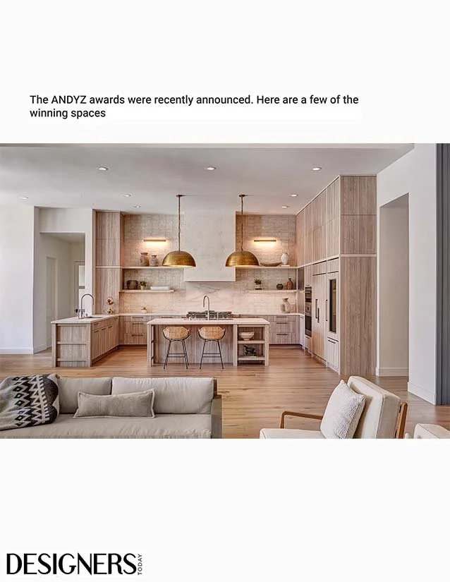Modern kitchen with wooden cabinets, two gold pendant lights, a kitchen island with two barstools, and open storage shelves, adjacent to a living room with a beige sofa and an armchair.