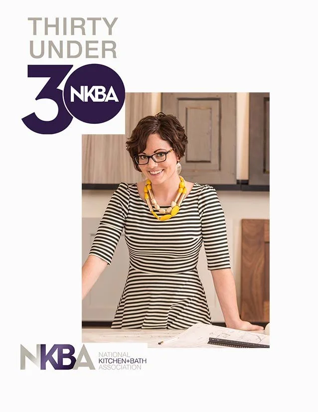 A woman wearing glasses and a striped dress with a yellow necklace, smiling and standing in a kitchen with cabinets, posing for a photo for the National Kitchen and Bath Association's 30 Under 30.