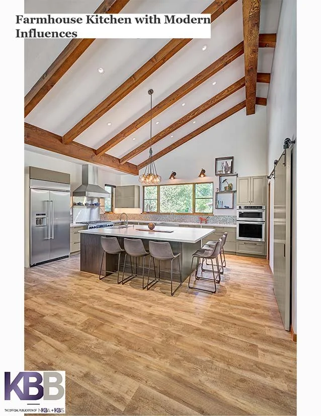 A spacious farmhouse kitchen with a vaulted ceiling featuring exposed wooden beams, a large white kitchen island with seating, stainless steel refrigerator, double oven, and ample white cabinetry. Large window above the sink allows natural light to f