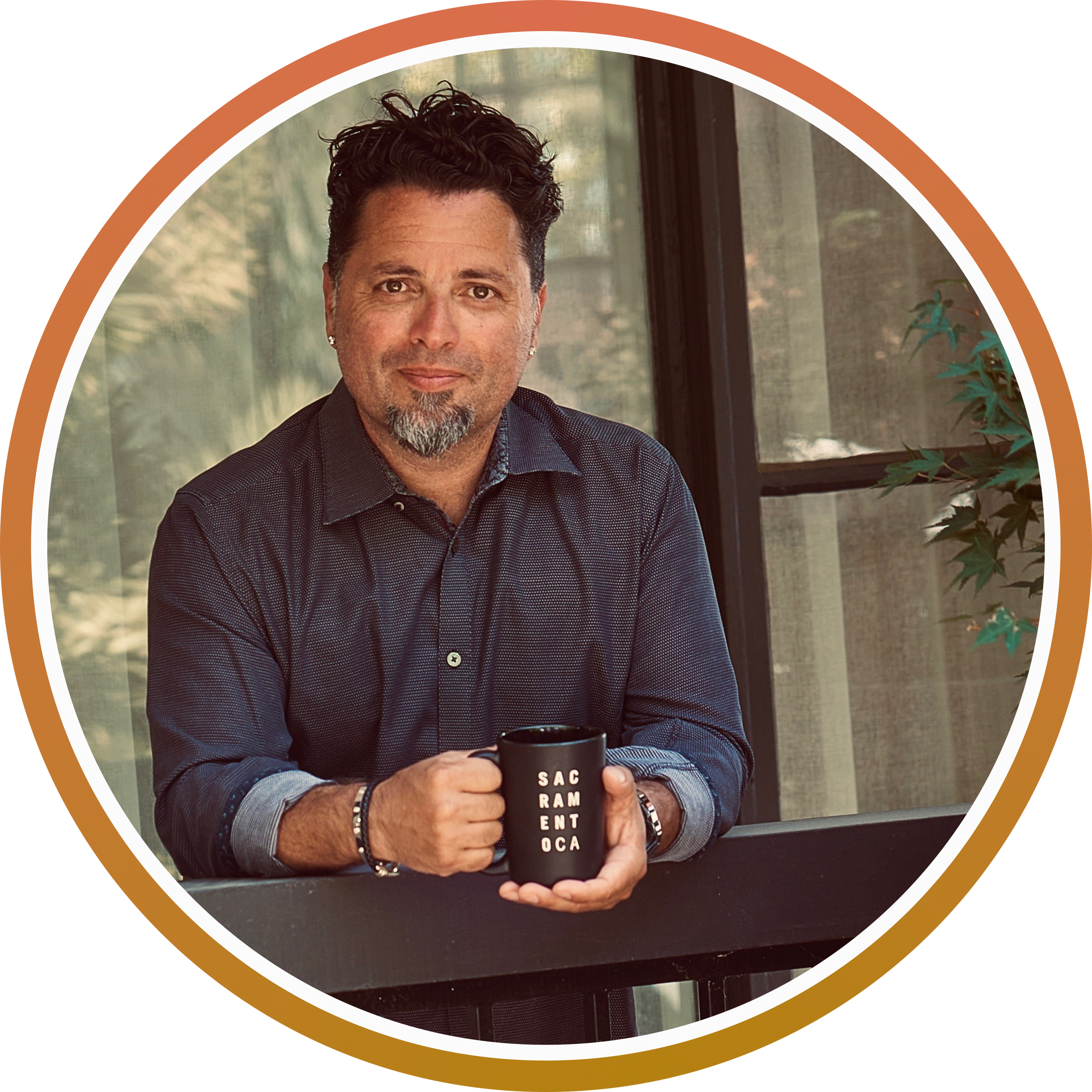 A man with dark curly hair, a goatee, and earrings holding a black mug with white letters that spell out 'SACRAMENTO' in a room with large windows and green plants.