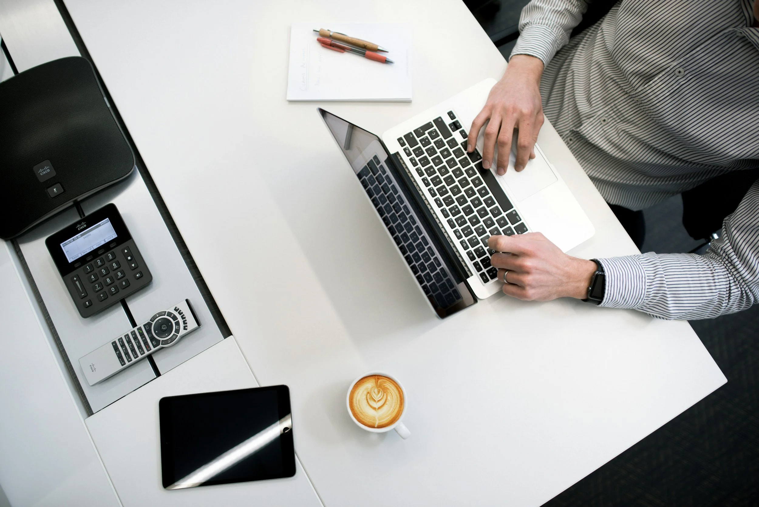 Overhead view of a person working on a laptop at a white desk with a smartphone, a remote control, a conference call speakerphone, a notebook with pens, and a cup of latte with latte art.