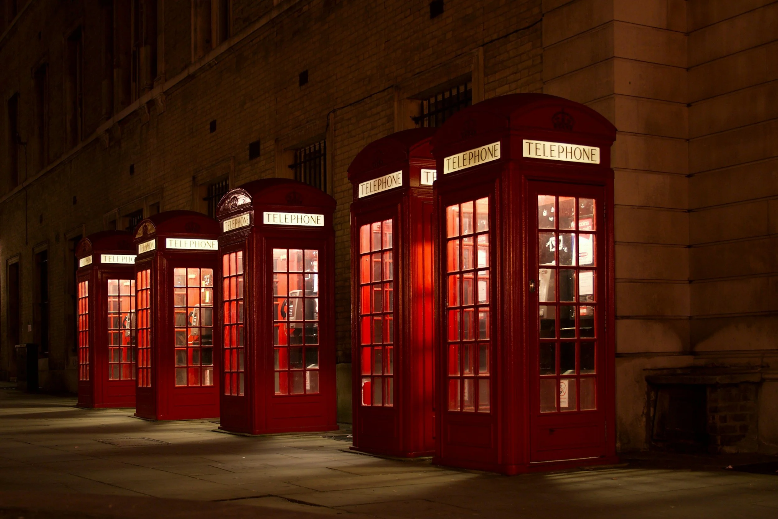Four red vintage British telephone booths lined up on a sidewalk at night, with warm interior lighting illuminating through glass panes, against a brick building wall.