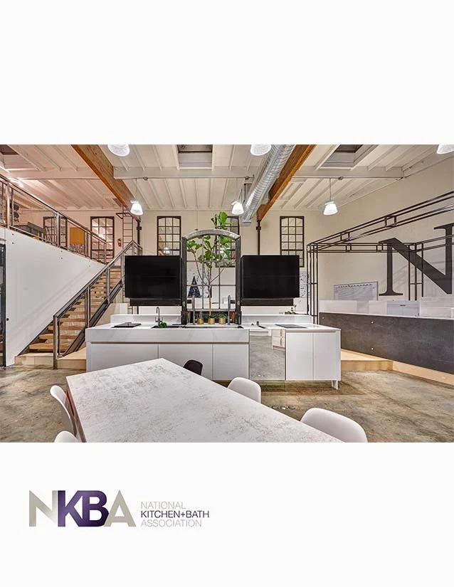 Modern kitchen space with white cabinets, two large black monitors, a table with white chairs, and industrial-style ceiling with exposed pipes, part of a workspace for the National Kitchen + Bath Association.