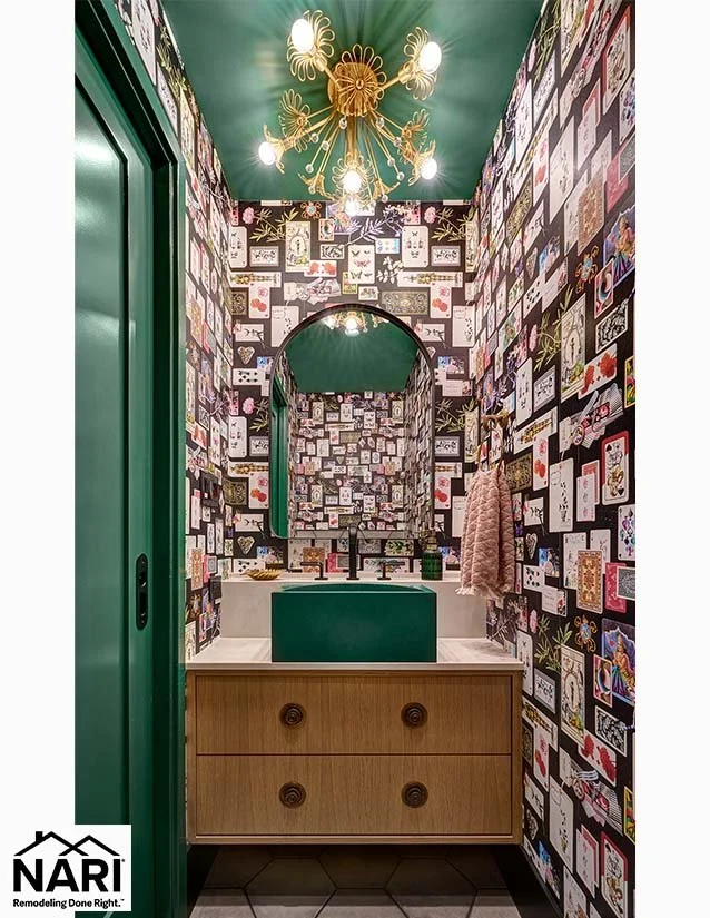 A small bathroom with wallpaper covered in various card and illustration designs. It features a green ceiling, a gold chandelier with multiple lights, and a modern wooden vanity. A pink towel hangs on the wall, and a large mirror is mounted above the