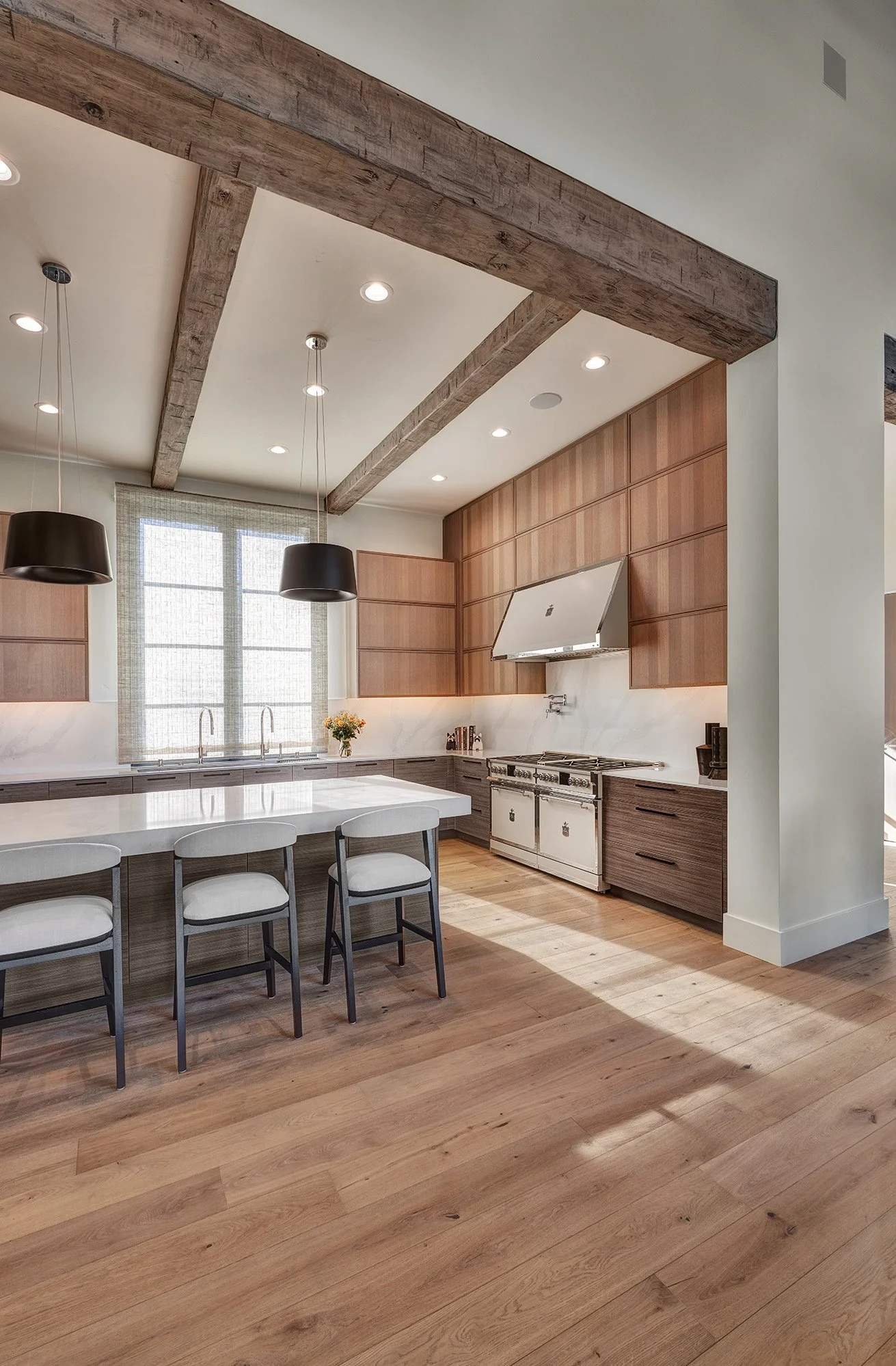 Modern kitchen with wooden cabinetry, a large kitchen island with white countertop, pendant lights, a window with semi-transparent blinds, hardwood floor, and an off-white range hood over a vintage style stove.