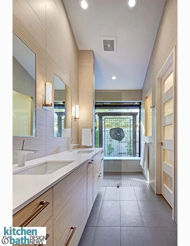 Modern bathroom with twin sinks, light wood cabinetry, large mirrors, and a bathtub at the far end with a decorative stone on a wire mesh in front of a large window.