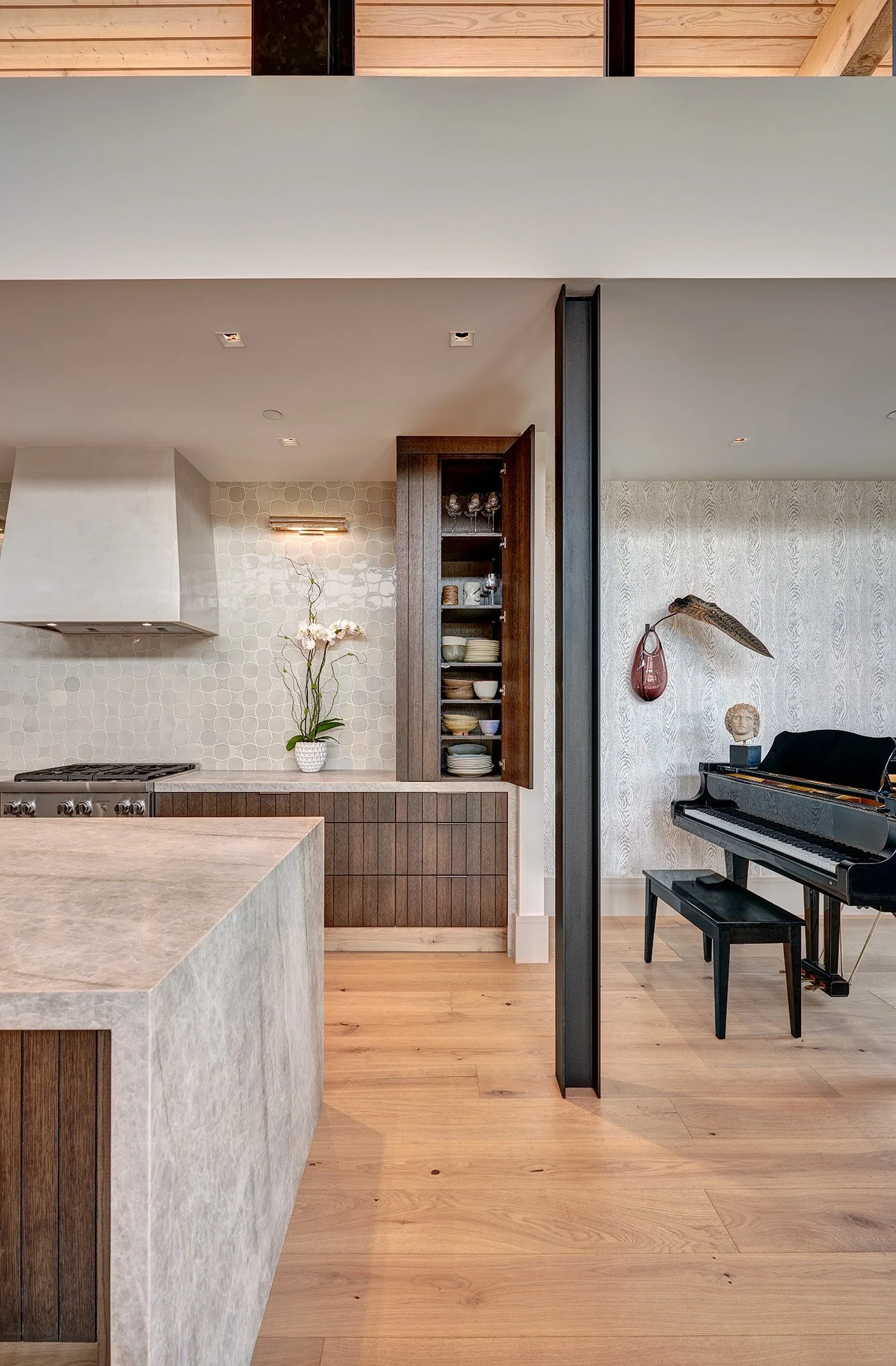 Interior of a modern home featuring a kitchen with wood accents, an orchid on the countertop, and a living area with a black grand piano, a decorative hanging vase, and a bust sculpture against light-colored walls.