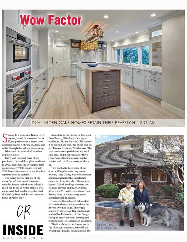 Interior view of a modern kitchen with white cabinets, a kitchen island with a gray base, built-in double ovens, and large windows showing outdoor greenery.