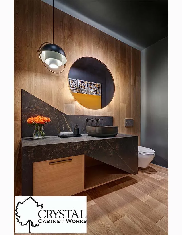 Modern bathroom with a wooden wall, a black stone countertop with a vessel sink, a round mirror, a wall-mounted light fixture, a toilet, and a small bouquet of orange flowers in a glass vase.