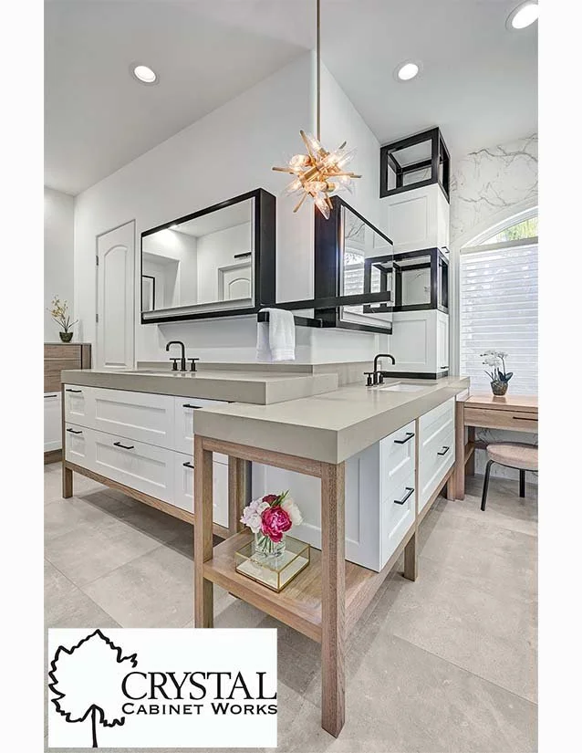 Modern kitchen with white cabinets, black accents, a large mirror, a dual sink, a wooden base island with a flower vase, and a dining area in the background.